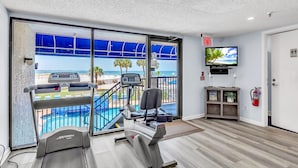 Ein Fitnessraum mit Laufbändern, Trainingsgeräten, einem Fernseher sowie Blick auf Pool und Strand.