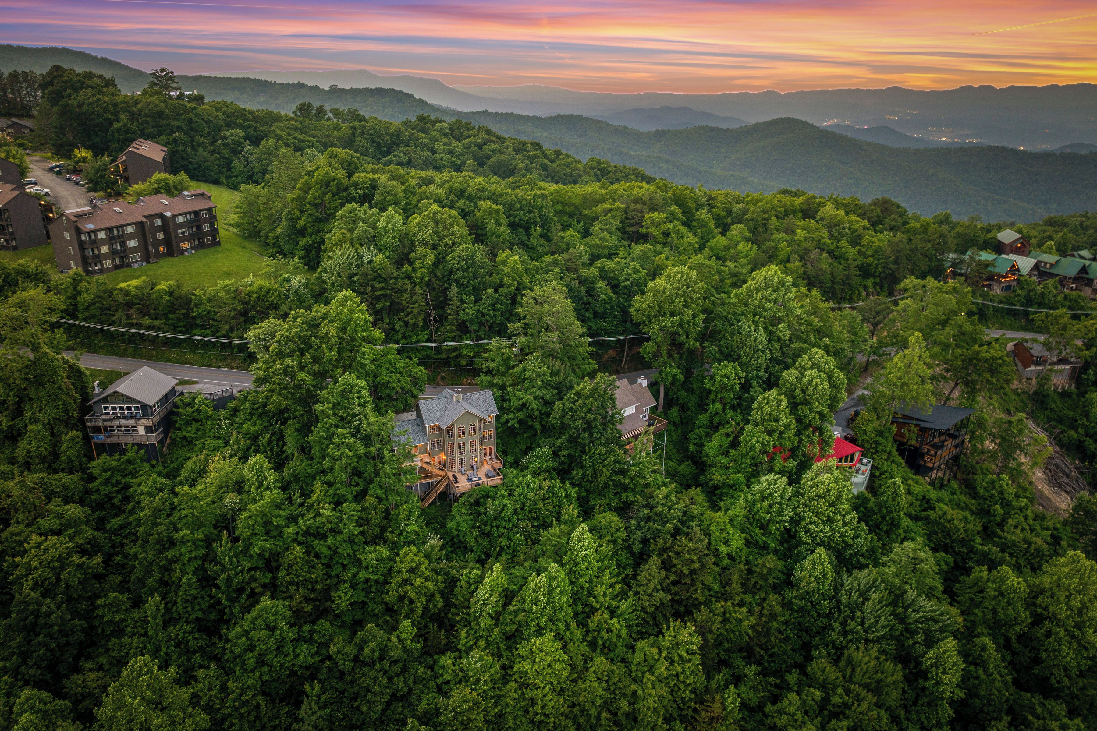 An aerial view of a residential area with multiple houses surrounded by dense forest, featuring a prominent house with a large deck and a smaller house with a red roof.