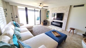 A modern living room with a sofa, ottoman, and a flat-screen TV mounted on a brick wall. - Sand Dollar 101 3 Bedroom Condo by RedAwning (St. Augustine)
