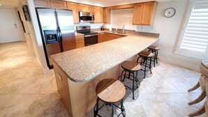 A modern kitchen with granite countertops, stainless steel appliances, wooden cabinets, and bar stools. - Sand Dollar 101 3 Bedroom Condo by RedAwning (St. Augustine)