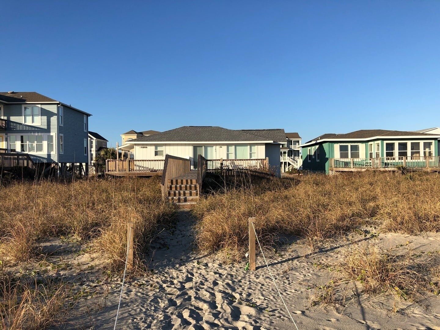 Une propriété située en bord de mer, avec une passerelle en bois menant à une maison, le tout entouré de hautes herbes et de sable.