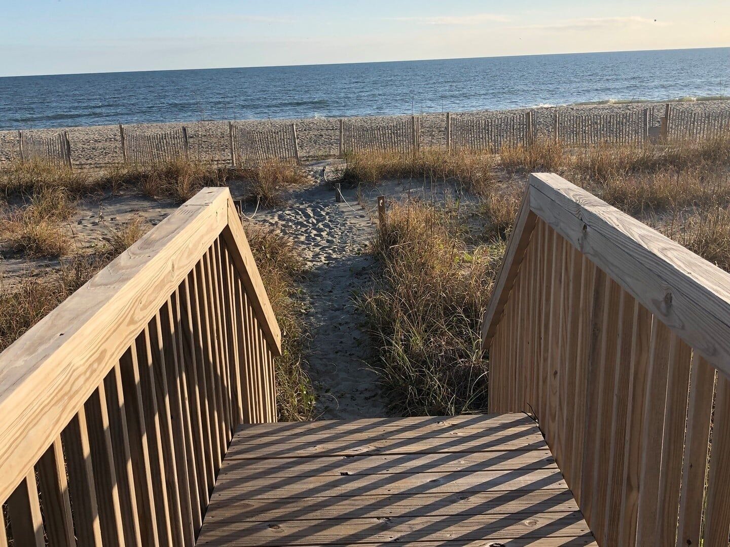 Une promenade en bois mène à une plage, avec un sentier sablonneux et une clôture en bois en arrière-plan.