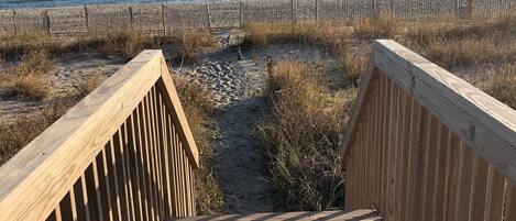 Une promenade en bois mène à une plage, avec un sentier sablonneux et une clôture en bois en arrière-plan.
