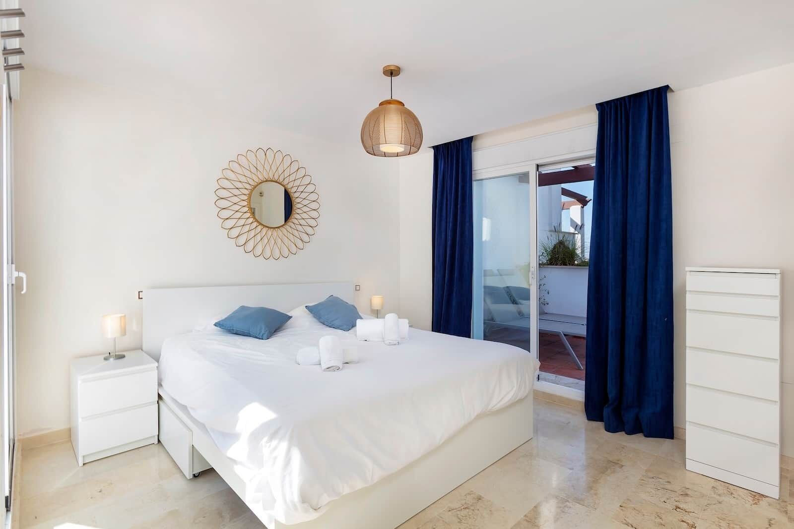 A modern bedroom with a large bed, a round mirror, a hanging light fixture, and a view through a glass door.