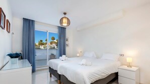 A modern hotel room with a large bed, two bedside tables, a glass table with a vase, and a view of palm trees through the balcony. - RDR230 - Apt. Aloha Royal (Marbella)
