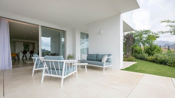 A modern outdoor living area with a sofa and chairs, a glass door leading to an interior space, and a view of a garden and mountains. - RDR233 - Apt. Palo Alto (Ojén)