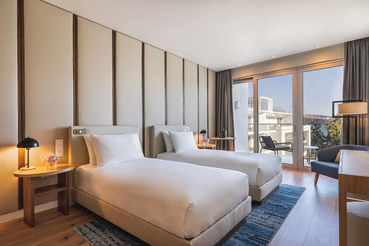 A hotel room with two beds, a desk, a chair, and a view of a building through large windows.