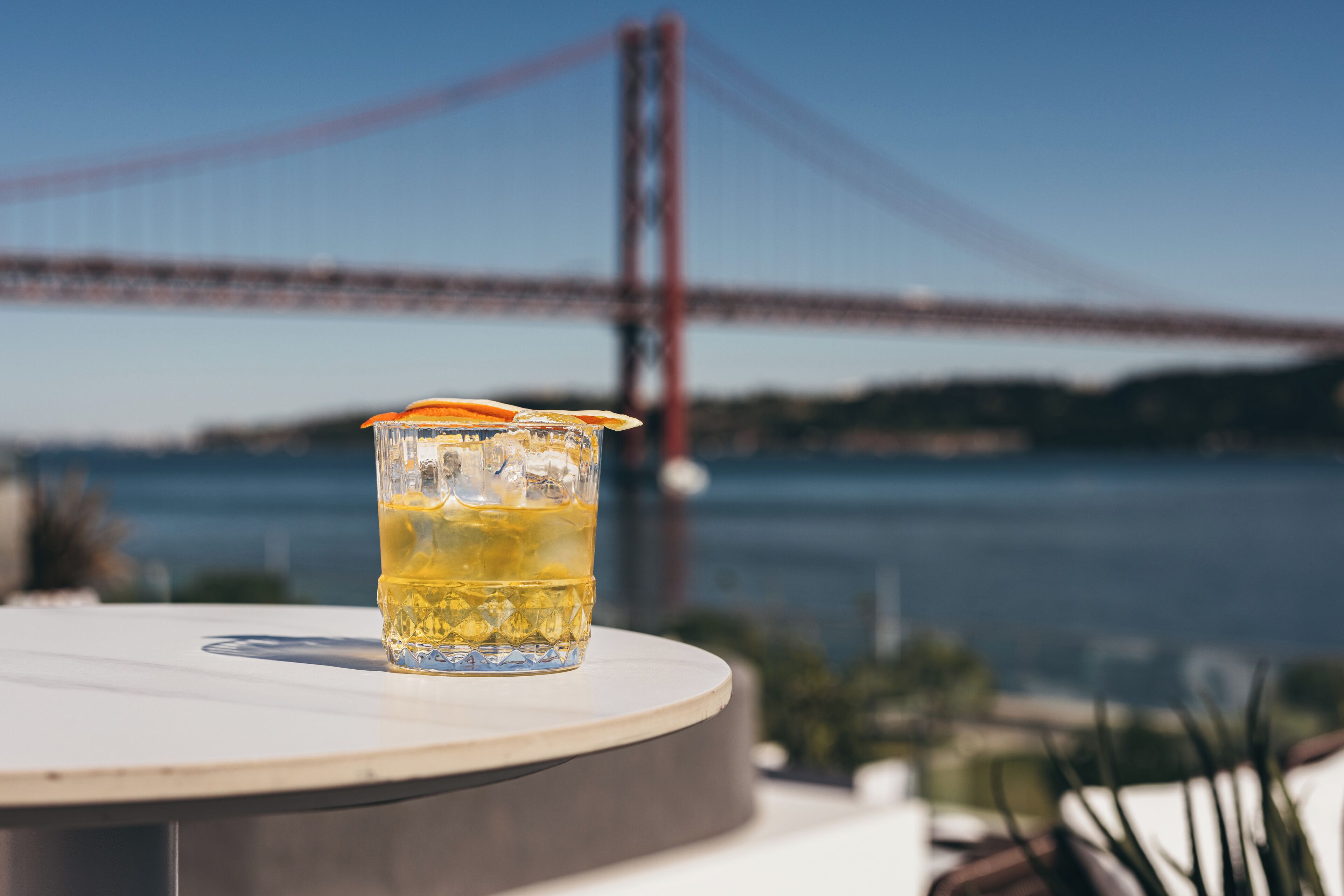 A glass of drink on a table with a view of a bridge in the background.