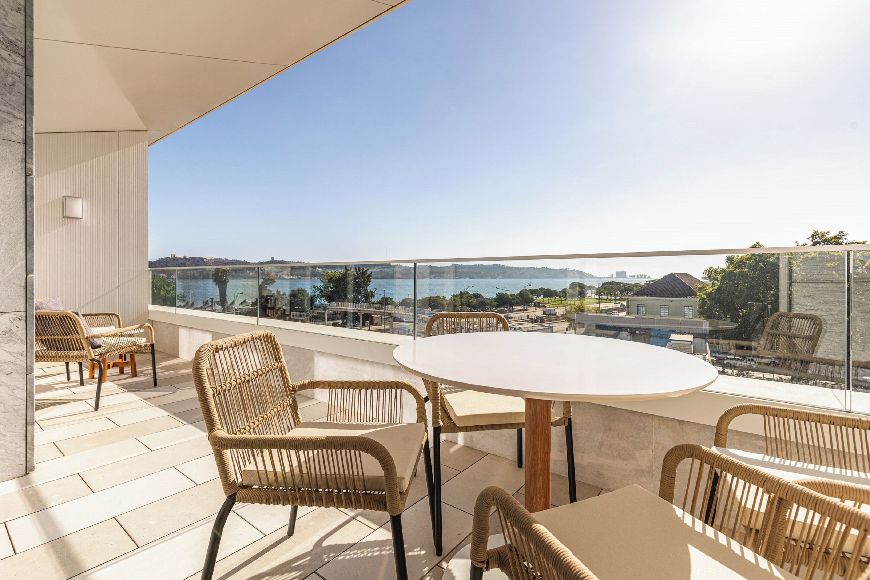 A balcony with wicker chairs and a round table, overlooking a body of water and distant buildings.