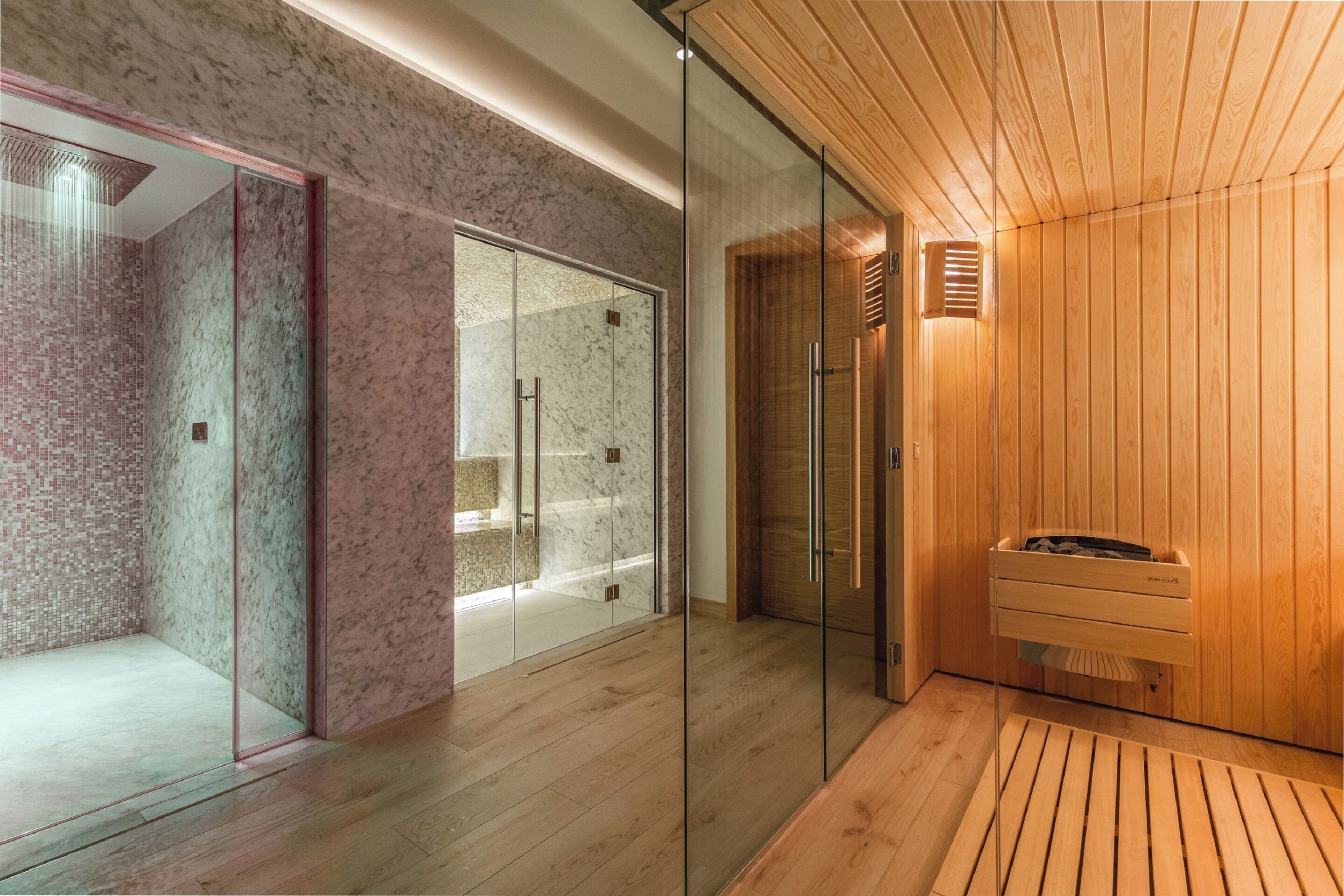 A modern spa room with a glass shower enclosure, wooden flooring, and a wooden sauna area.
