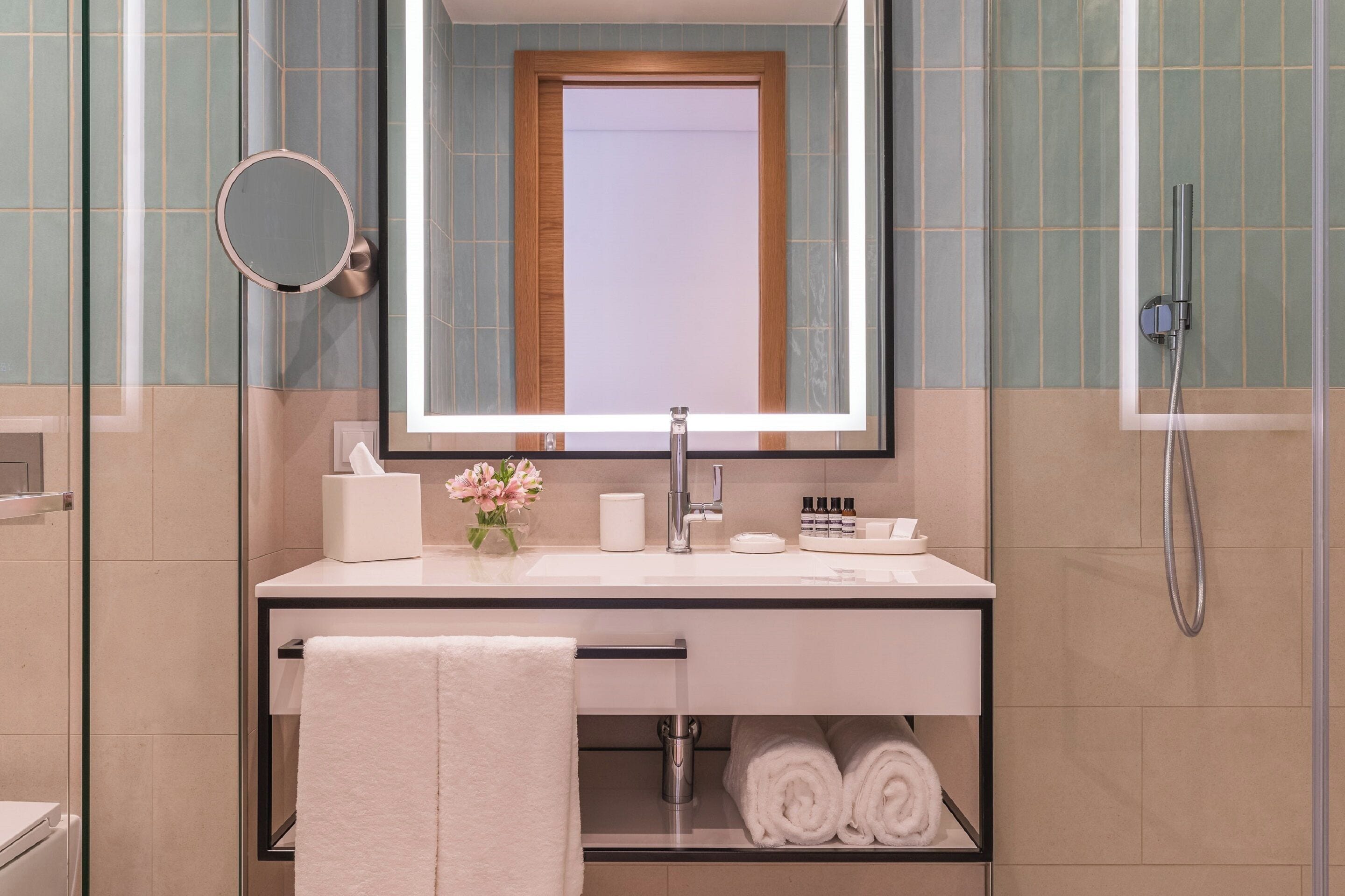 A modern bathroom with a large mirror, a sink with toiletries, a towel rack, and a shower area.