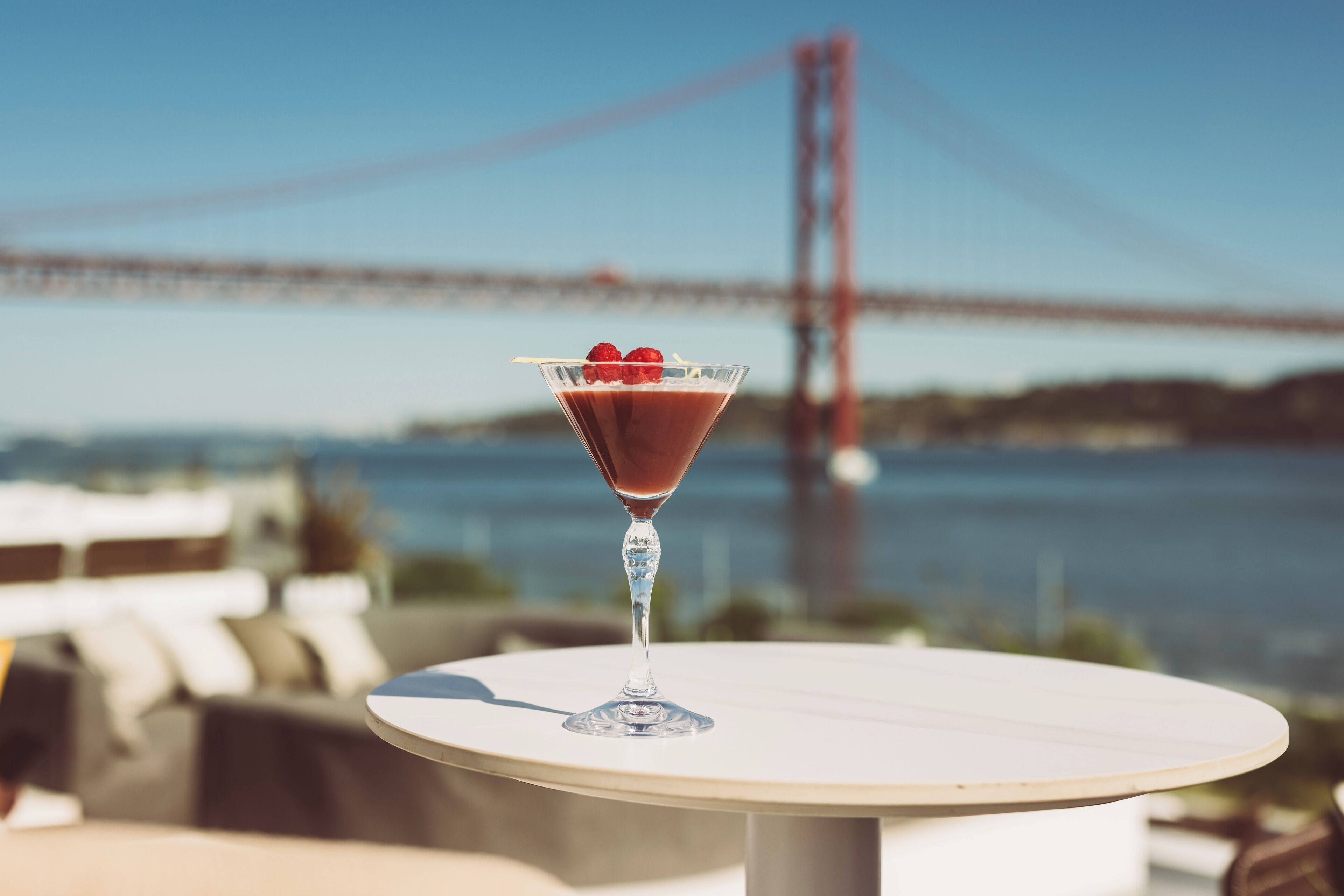 A martini glass with a red drink and raspberries on a round table with a view of a bridge in the background.