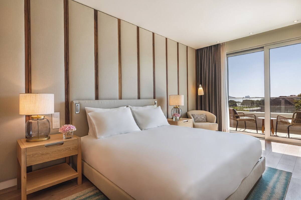 A hotel room with a large bed, two bedside tables with lamps, a small table with a vase of flowers, a chair, and a balcony with a view.