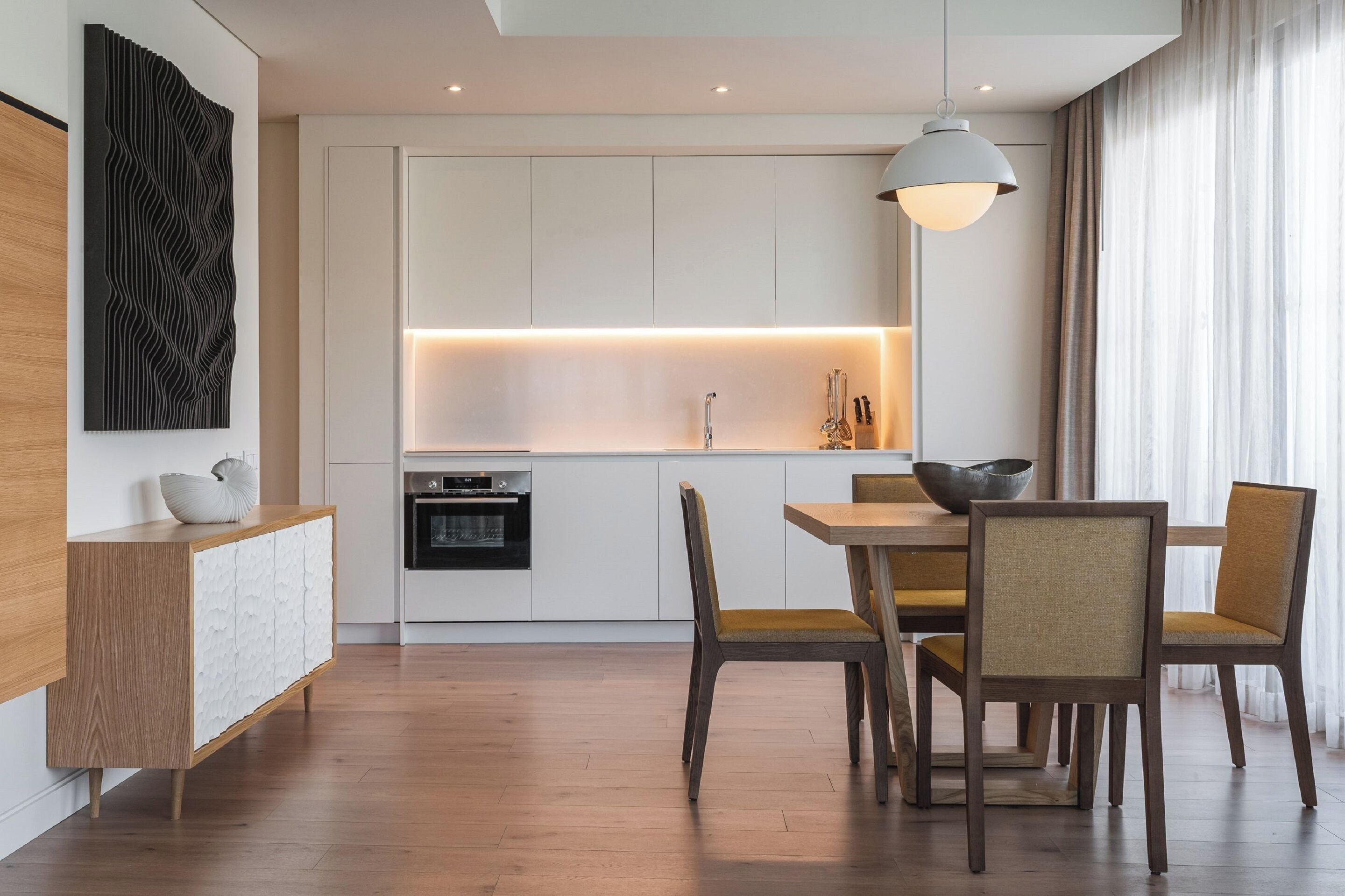 A modern kitchen with white cabinetry, a wooden dining table, and chairs. There is a pendant light above the table and a decorative wall piece on the left.