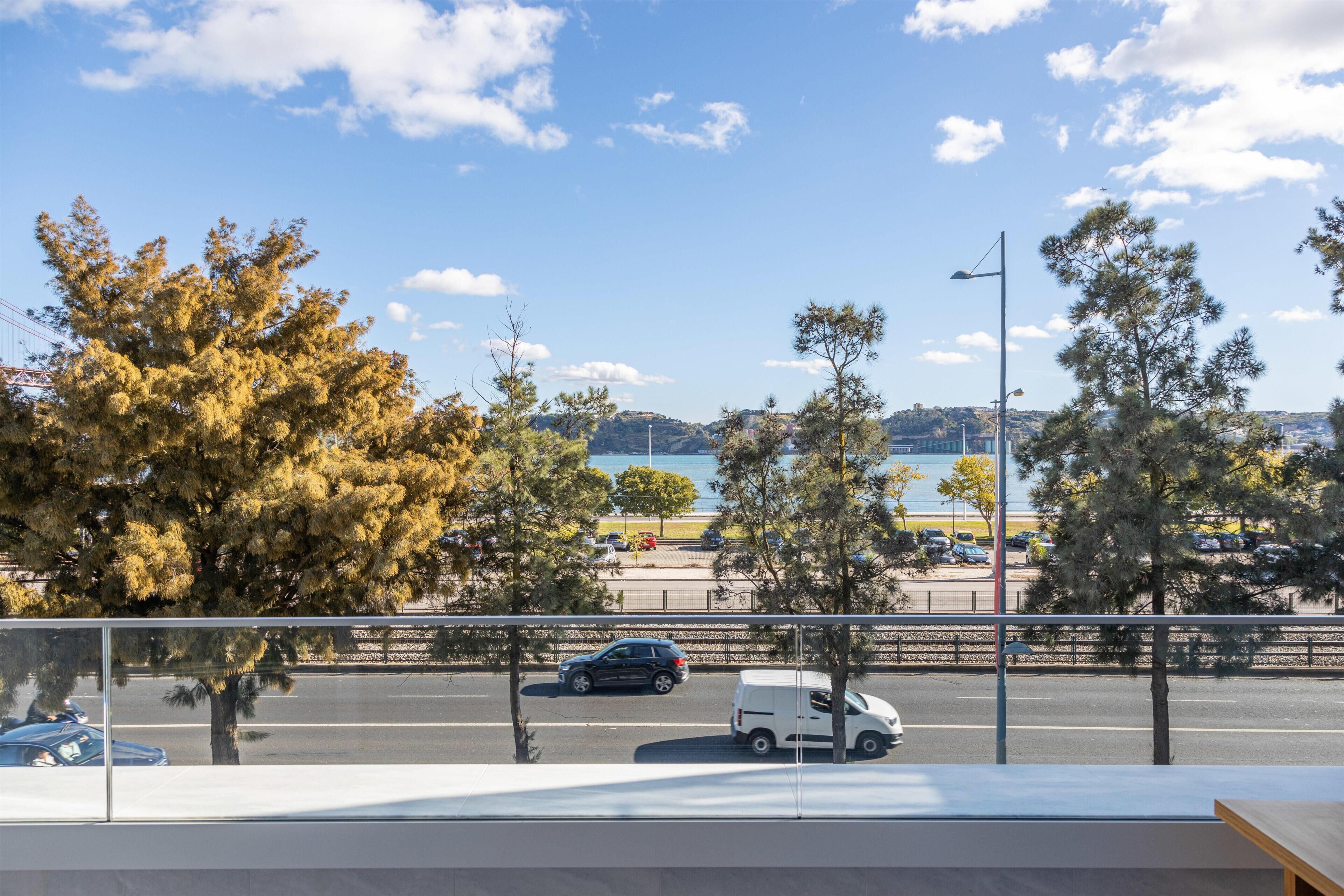 Une vue depuis un balcon donnant sur un parking bordé d’arbres, avec une étendue d’eau visible au loin.
