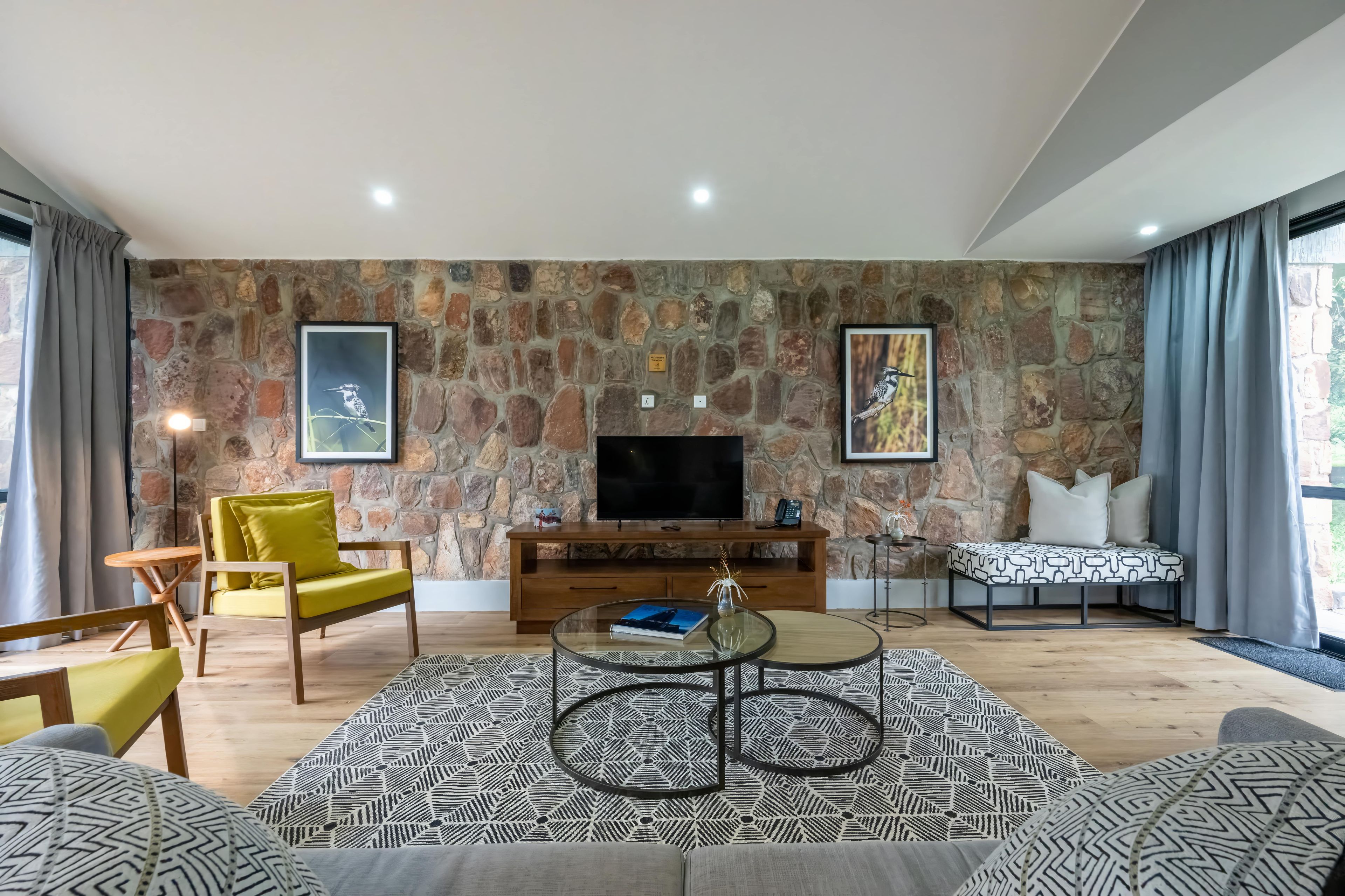 Uma sala de estar moderna com parede de pedra, TV de tela plana, poltrona amarela e tapetes estampados.
