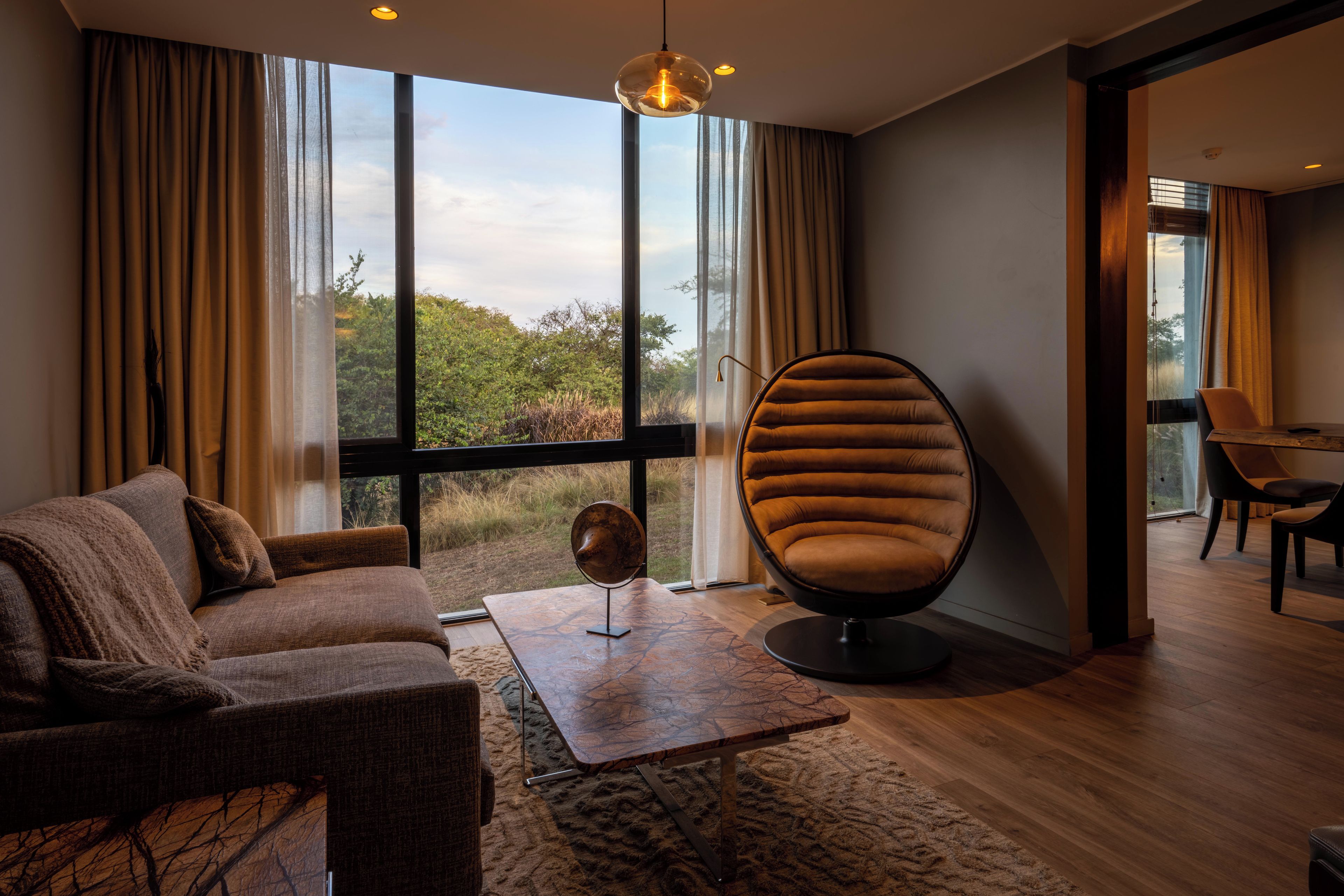 A modern living room with a sofa, a unique egg-shaped chair, a coffee table, and a view of the outdoors through large windows.