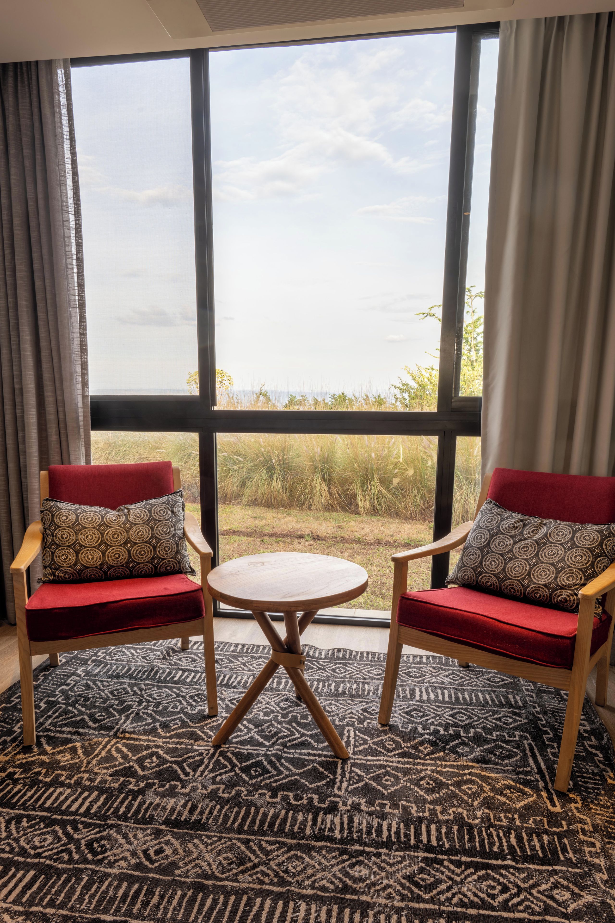 Una habitación con dos sillones tapizados en rojo, una mesa redonda pequeña y un ventanal con vistas al exterior.
