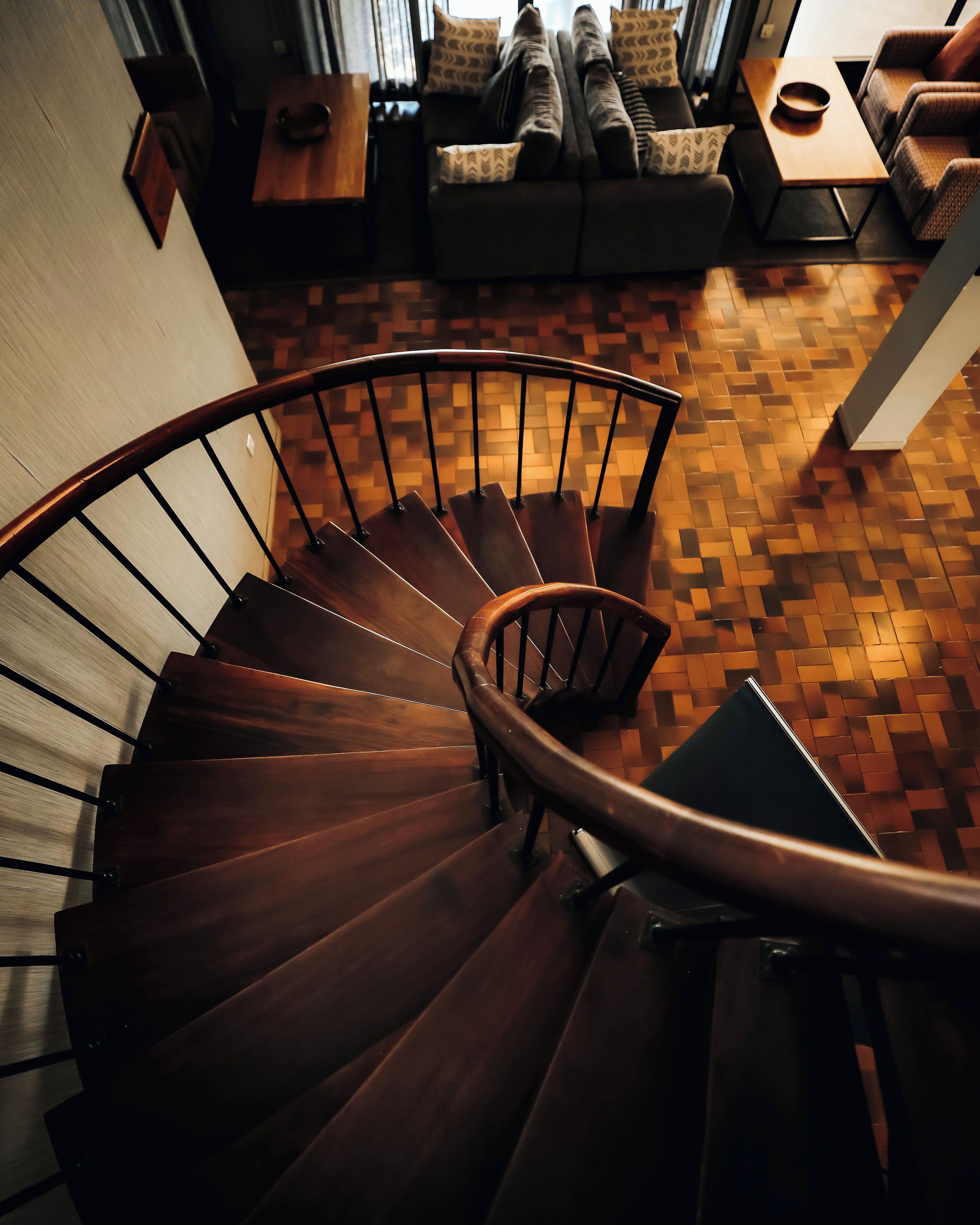 A wooden spiral staircase with a handrail, leading to a living area with a patterned floor and furniture.