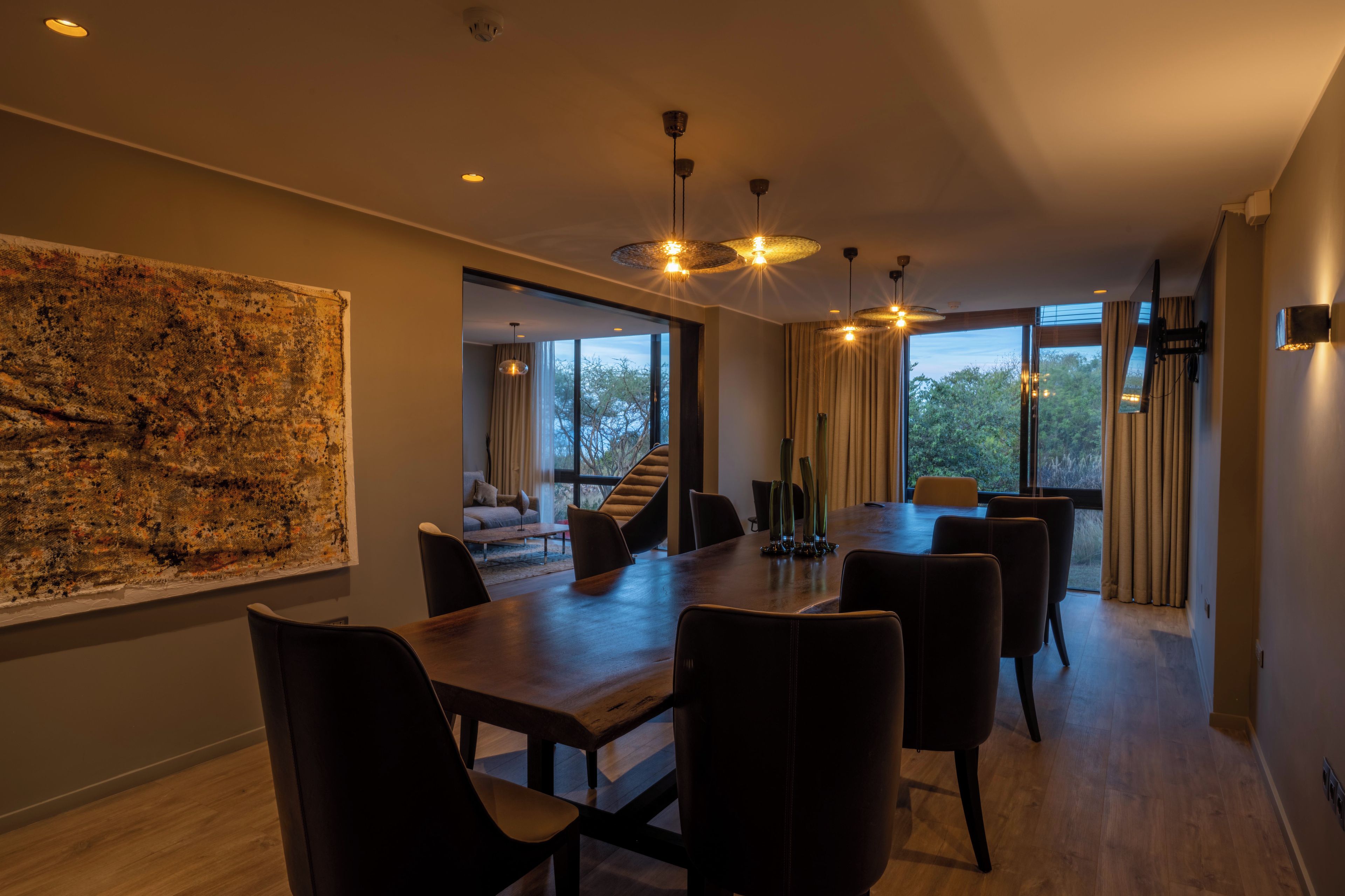 A modern dining area with a large table and chairs, a view of the outdoors through large windows, and a large abstract painting on the wall.