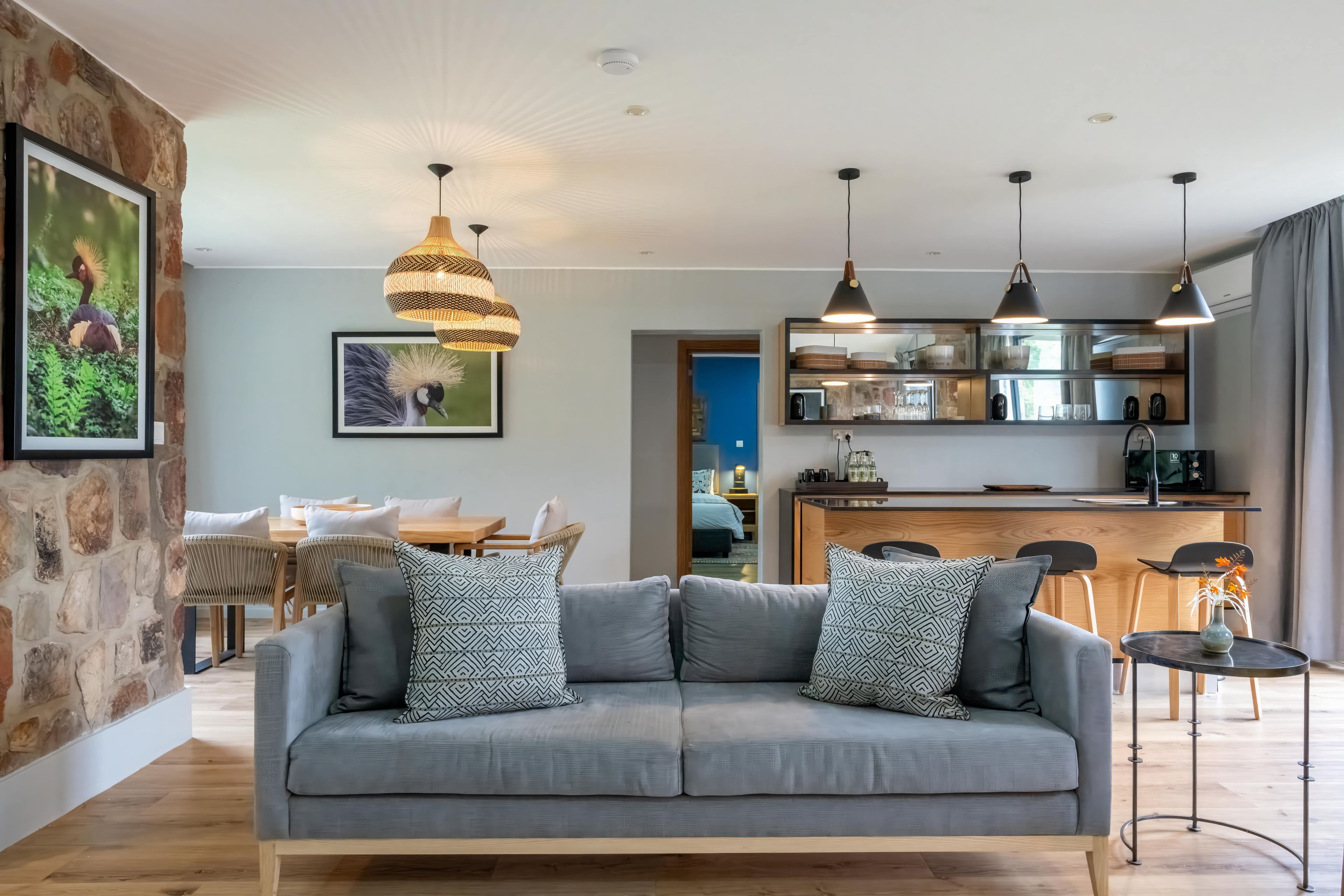 Una sala de estar moderna con un sofá gris, cojines estampados, una zona de comedor con mesa y sillas de madera, y una cocina con isla integrada e iluminación colgante.