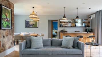 Una sala de estar moderna con un sofá gris, cojines estampados, una zona de comedor con mesa y sillas de madera, y una cocina con isla integrada e iluminación colgante.
