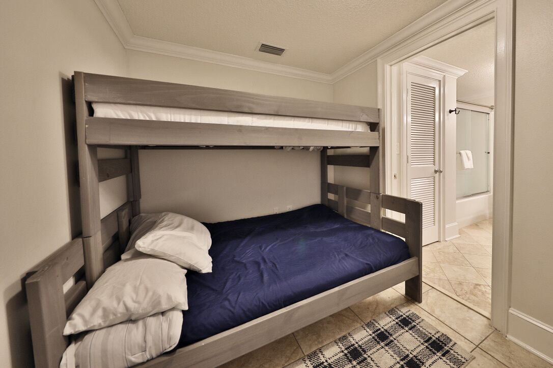 Ein Stockbett mit blauer Matratze und weißen Kissen, ein karierter Teppich und eine Tür zu einem Badezimmer mit Dusche.