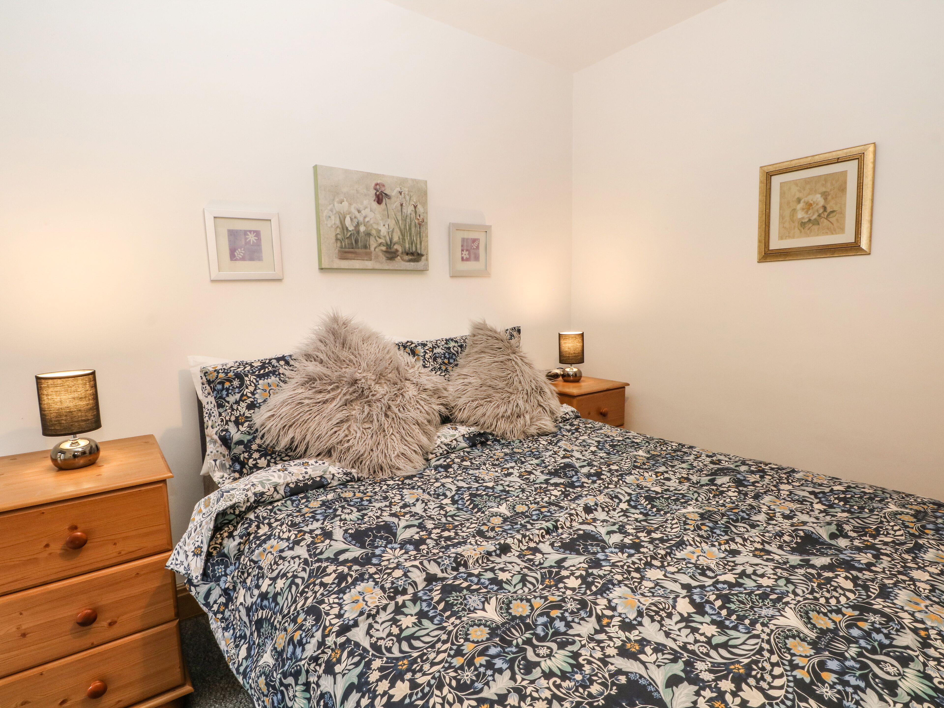 A bedroom with a floral-patterned bedspread, two fluffy pillows, a wooden nightstand with a lamp, and three framed artworks on the wall.