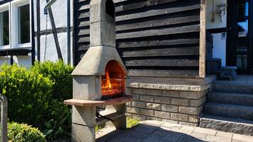 Un four extérieur en pierre, avec un feu allumé à l’intérieur, installé sur une surface pavée, avec une structure en bois en arrière-plan.