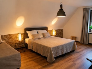 A bedroom with a bed, two bedside lamps, a wooden headboard, a small stool, and a window with curtains.