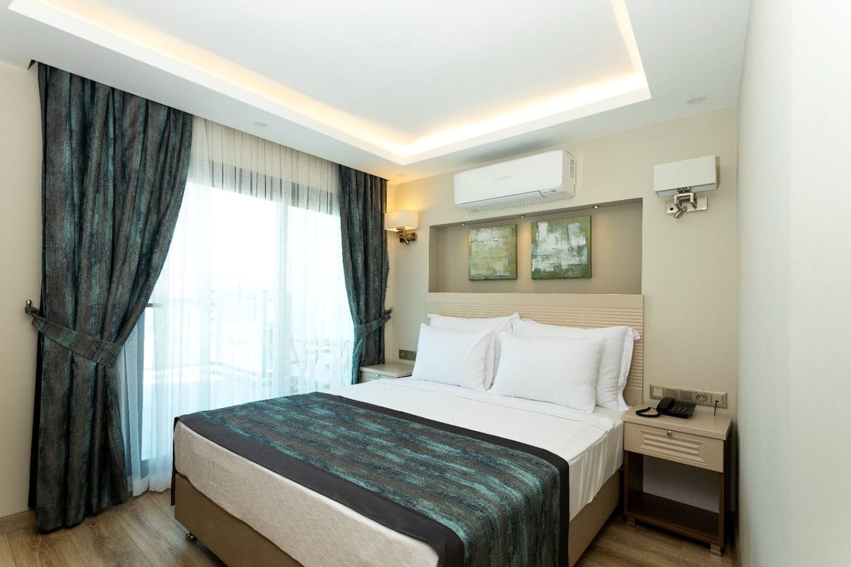 Een hotelkamer met een bed, gordijnen, een airconditioningapparaat, een nachtkastje met een telefoon en kunst aan de muur.
