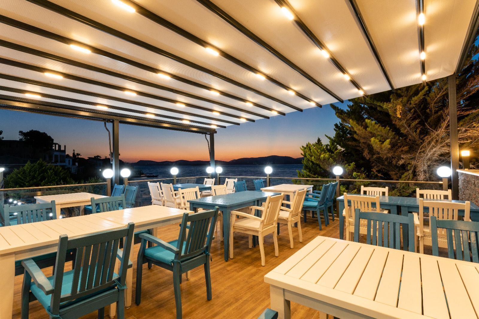 Un espace extérieur aménagé pour les repas, avec des tables et des chaises, une pergola illuminée et une vue sur la mer au crépuscule.