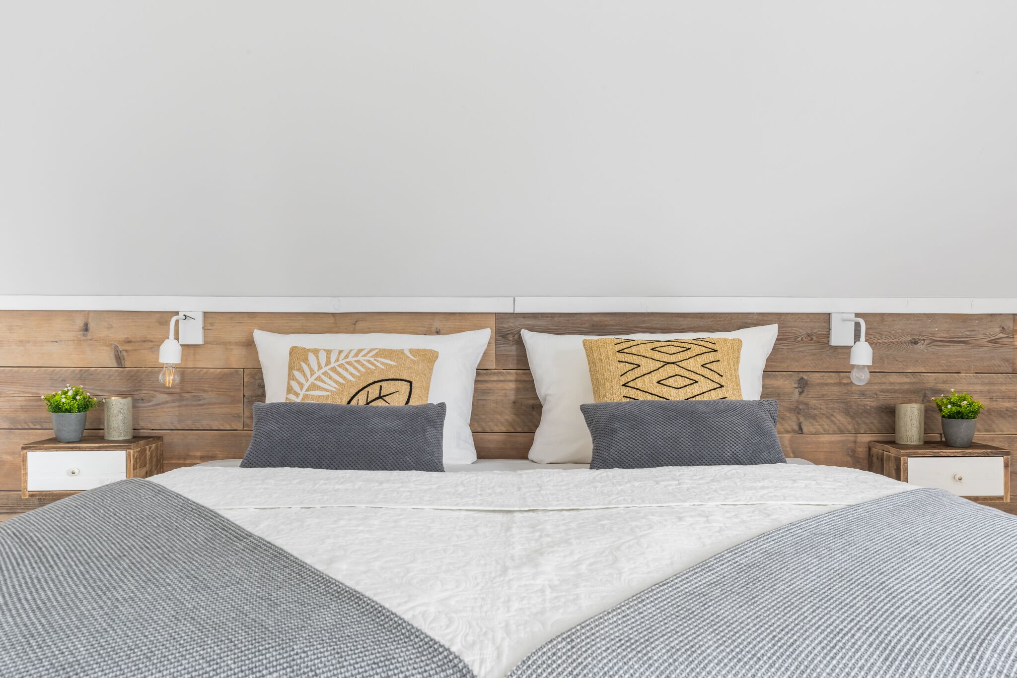 Een netjes opgemaakt bed met een wit-grijze deken, twee kussens met geometrische patronen en een houten hoofdbord.