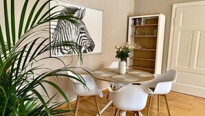 A dining area with a round table, white chairs, a zebra artwork on the wall, a bookshelf, and a potted plant.