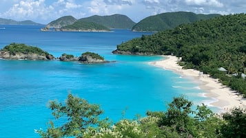 A coastal landscape with clear blue waters, a sandy beach, and lush green hills in the background.