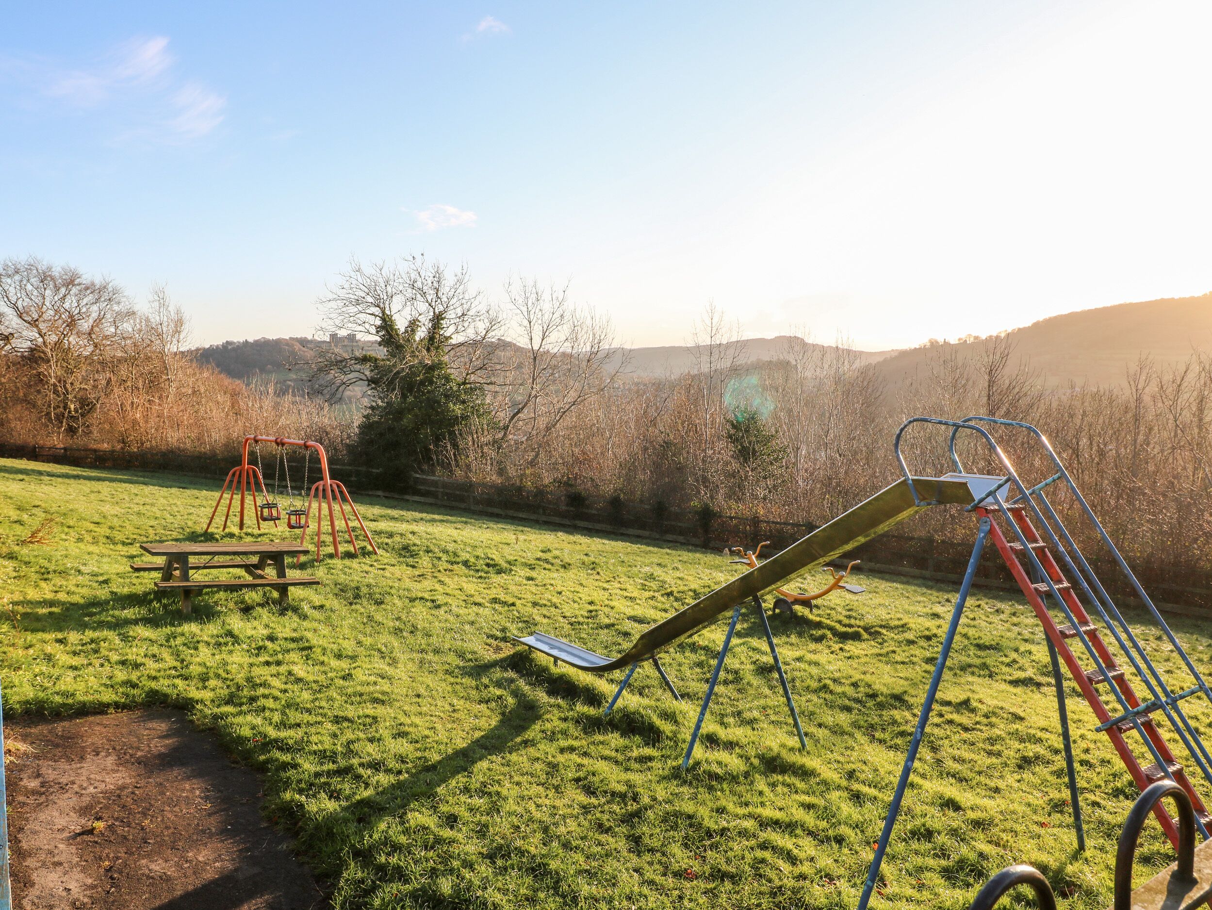 A playground with swings and a slide in a grassy park area with trees and hills in the background.