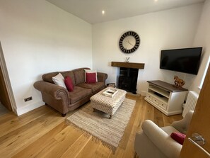 A living room with a brown sofa, a white armchair, a coffee table, a fireplace, a TV, and a clock on the wall. - Cavendish Cottage (Matlock)