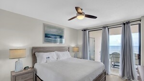 Un dormitorio con cama, mesitas de noche, ventilador de techo, un cuadro y un balcón con cortinas.