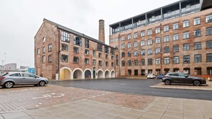 A parking area with two cars, a brick building with multiple windows, and a modern structure with balconies. - Modern 2 Bedroom Riverside Apartment in Leeds (Leeds)