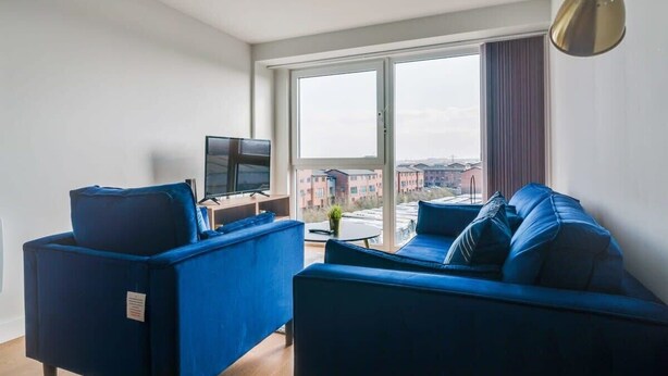 A modern living room with a blue sofa set, a flat-screen TV, and a large window offering a city view. - Modern 2 Bedroom Riverside Apartment in Leeds (Leeds)