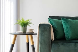 A room with a green sofa, a small table with a potted plant and a cup, and a window with blinds.