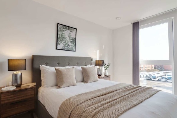 A hotel room with a large bed, two bedside tables with lamps, a framed artwork above the bed, and a view of parked cars through a large window.