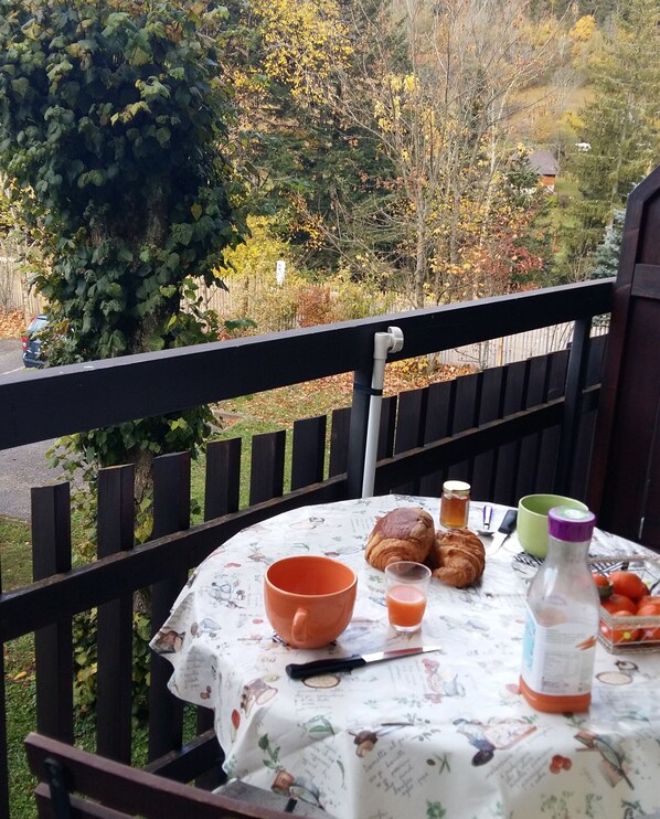 Une table ronde recouverte d’une nappe à motifs floraux, un croissant, un verre de jus d’orange, une bouteille et un bol, le tout placé sur un balcon d’où l’on aperçoit des arbres et une clôture.