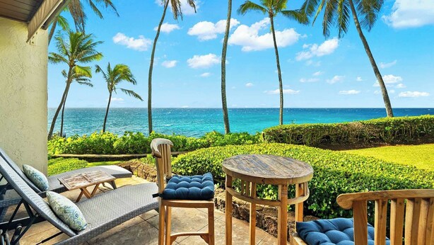 Eine Terrasse mit Korbmöbeln, einem Holztisch und Blick auf das Meer und Palmen.