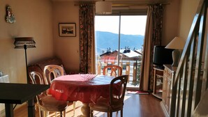 Un comedor con una mesa dispuesta para cuatro, un televisor sobre un mueble y vistas a montañas a través de un ventanal grande.
