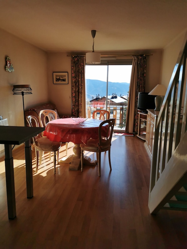 A dining area with a table set for four, a television on a stand, and a view of mountains through a large window.