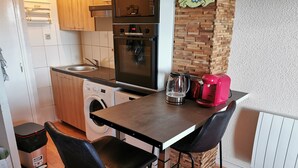 Cocina compacta con lavadora, cafetera y hervidor sobre la encimera. Hay dos sillas negras y una mesa pequeña.