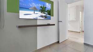 Moderni hotellihuone, jossa on suuri, seinälle asennettu litteänäyttöinen televisio. Sen alla on puinen hylly, ja huoneesta on ovi toiseen huoneeseen.