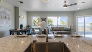 A modern kitchen with a large island, a dining area with a table and chairs, a sofa, and a balcony with a view of the beach. - Casa Caribe-Soundside Private Home w/Pool and Tennis 2 Min from the Beach (Navarre)