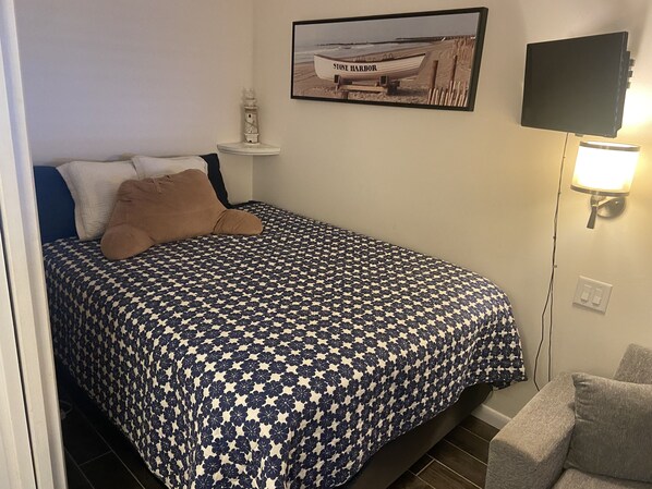 Un dormitorio con una cama, una mesita de noche, un televisor de montaje en pared y un cuadro con una imagen de un barco.